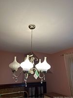 Wide shot showing the entire chandelier hanging from ceiling with five white glass shades and brass frame with crystal drops.