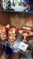 Photo showing the copper plate and six copper cups on a glass shelf in a wooden cabinet.