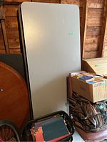 Single photo showing a gray folding table in folded vertical position leaning against a wooden wall. Nearby items such as boxes and a bicycle wheel partially visible.