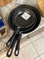 Three nested black frying pans with black handles. The pans appear to have non-stick interiors and look new inside.