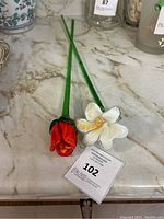 Two glass flowers placed on a marble surface, one red and one white, showing long green stems and detailed petals.