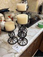 Set of three black metal twisted design votive candle holders with cream-colored pillar candles arranged on a kitchen counter with vintage containers in background.
