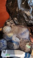 Mesh bag filled with approximately 12 assorted soccer balls in various colors including white with blue, black, and yellow marks. Some balls show signs of wear.