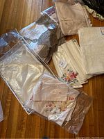 Various folded and packaged table linens including embroidered napkins and tablecloths, some in plastic bags, with floral and geometric designs in white, cream, and pink colors.