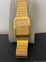 Front view of the ADEC gold tone watch on a grey display cushion showing the square face and metal link bracelet.