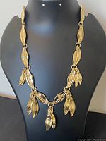 Front view of gold-tone leaf design necklace arranged on a black display stand showing the shape and detail of leaf links and dangling leaf charms.