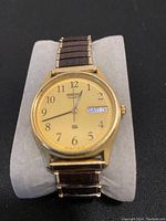 Front view of Seiko gold tone quartz watch on soft stand, showing beige dial with Arabic numerals and day-date window.