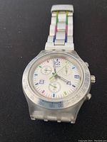 Front view of Swatch chronograph wristwatch with multicolor metal bracelet and clear plastic case resting on a black surface.