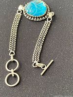 Close-up front view showing blue carved themed stone centerpiece and sterling silver box chain sides with toggle clasp open.