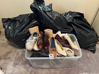 Three large black bags and one clear plastic tub containing assorted used women's shoes in a variety of styles and conditions.