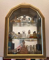Full view of gold-framed mirror cabinet with three glass shelves holding assorted perfume and toiletry bottles.