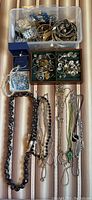 Full lot of assorted costume jewelry displayed in plastic box and laid out on surface including necklaces, bracelets, earrings, and pins.