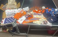 Photo showing a variety of Denver Broncos collectibles including knit sweater, scarves, stuffed bears, drinking glasses, and vintage newspapers laid out on a table.