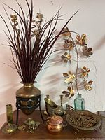 Wide view of lot including brass vase with red artificial foliage, brass floral wall sculpture in glass bottle, hammered copper bowl with brass butterfly, and various small brass and copper items on wooden surface.