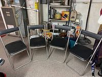 Four mid-century folding chairs with chrome frames and black vinyl seats and backrests, arranged side by side indoors in a basement setting with various household items.