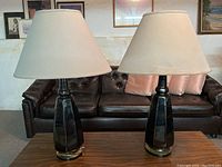 Pair of mid-century style ceramic lamps on a wooden coffee table in a vintage-style room. Both lamps have dark faceted bases and large, tapered fabric shades that are showing water damage.