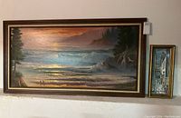 Two framed paintings displayed against a wall, a large seascape and a smaller street scene painting to the right.