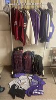 Wide view of rack with men’s XS/S clothing including shirts and jackets and luggage below; shows lot contents and general condition needing cleaning.