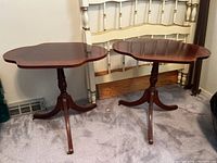 Pair of vintage wooden side tables with scalloped edges on tabletops, tripod pedestal bases, dark reddish wood finished, standing together in a carpeted room against a bed frame.