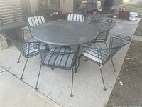 Full view of round wrought iron table surrounded by six wrought iron chairs with striped cushions, set on a concrete patio.