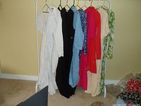 Photo of 7 dresses on hangers against wall, showing a range of colors and styles including white, black, blue, red, beige, and floral print