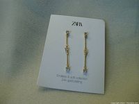 Photo of one pair of ZARA earrings attached to branded card, showing long drop design with 24k gold plating and crystal highlights.