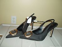 Pair of black slingback high heel shoes shown from side angle, highlighting pointed toe and gold buckle detail with original tag still attached