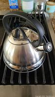 Full view of stainless steel kettle with black handle on stove grate showing its overall condition and design.