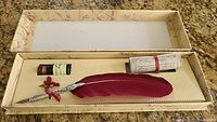 Full view of open box containing calligraphy kit items: red feather quill pen with pewter tip, ink bottle, and rolled instructions.