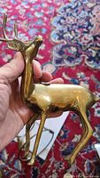 Photo of solid brass deer figurine held in hand showing side profile and antlers, placed on patterned carpet.