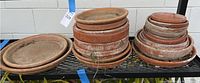 Wide view showing three stacks of terracotta clay plant saucers of various sizes arranged on metal shelving showing weathering and dirt.