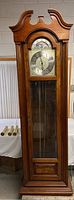 Full frontal view of the tall grandfather clock showing wooden case and brass clock face with glass door closed.