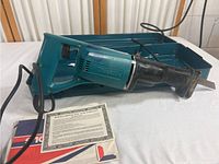 Makita reciprocating saw with power cord and instruction manuals, laid on a table with a blue carrying case in background. Includes extra blades.