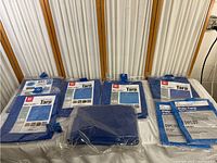 Photo of six new blue tarps in packaging, mostly 5x7 ft sizes with one much larger tarp.