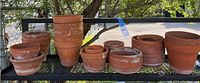 Wide shot of the entire collection showing 11 terra-cotta pots of varying sizes arranged outdoors on metal shelving with tree and fence background.