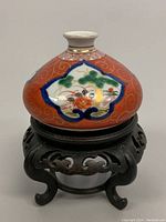 Front angle showing hand painted Kutani porcelain vase with floral and nature motifs on a red background and wooden carved base.