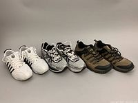 Three pairs of men's shoes shown side by side on plain background. From left to right: white K-SWISS sneakers with black stripes, silver FILA sneakers with black details, tan and black McKinley hiking shoes.