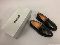 Photo showing the pair of black leather tassel loafers with their original white Unlisted box placed beside them on a white background.