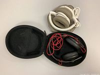 Two pairs of headphones shown: one in a black carrying case with red cable, the other white and brown with USB cable coiled and placed beside.