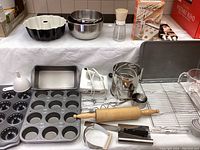 Wide angle image showing various baking pans, a wooden rolling pin, electric hand mixer, metal sifter, cooling racks, and other baking tools arranged on a white cloth.