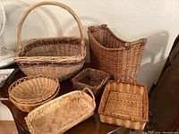 General photo showing six woven baskets of varying sizes and shapes, displayed on a wooden surface against a beige wall.