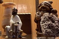 Photo showing carved wooden figurine of a smiling man with hat holding a lantern, small ceramic or porcelain robed man sitting on animal, and porous rock in front