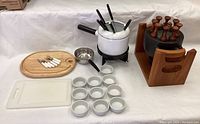 Full view of the fondue set pieces arranged on a table showing the white ceramic pot, wooden cutting board with knives, ceramic bowls, and black cast iron pot with wooden rack.