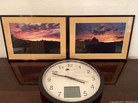 Two framed sunset photos side by side above a wall clock with digital thermometer.