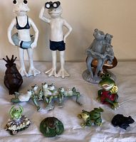 View of all frog figurines on a table including two large white frog statues, cast iron frog king, group of four lounging frogs, and other small frog figurines