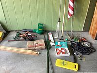 Full lot overview of all gardening tools and accessories laid on floor showing brush cutter, electric blower, rake, garden stakes, hummingbird feeder box, hose, pruners, paper lawn bags, and hand tools.