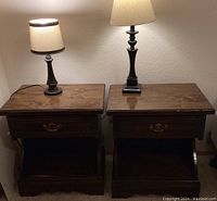 Two matching wooden night stands each with a drawer and an open shelf underneath, accompanied by two table lamps of different heights and shapes on top. The wood shows natural finish and color variations.