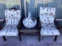 Front view of two rustic wooden patio chairs attached by a built-in table, with floral patterned cushions and a white ceramic basket-style planter on the table.