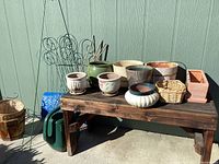Rustic wooden bench with various ceramic and wicker planter pots, metal plant stakes, watering can, and trellis arranged on and near it.