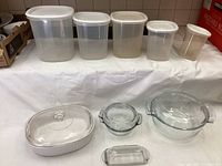 Five translucent Rubbermaid storage containers with white lids in varying sizes along the back of the photo. Arranged in front are two covered Pyrex baking dishes (oval with white ribbed base and round clear glass) and one Corningware white ceramic covered dish, along with a small clear glass butter dish with lid.